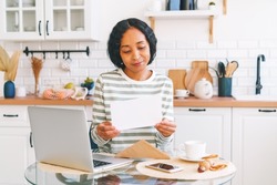 Smiling African-American woman reading paper letter in kitchen. Concept of receiving mail correspondence. Opening envelope. Postage hobby. Looking for news with enthusiasm