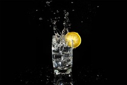 heavy splashing of an ice cube in a glass of fizzy drink decorated with a lemon. Splash on black background with reflection on the base. vertical format. Hard ligth