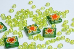 Green glass seed beads and millefiori flower beads scattered on a light colored background. Closeup, shallow depth of field.