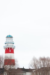 A tall red and white striped lighthouse. Several houses are on the bottom of the frame and snow melting on a ground, slightly showing it. White cloudy clear bright sky is above