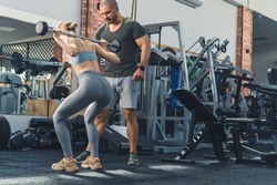 Back view of a young adult caucasian brunette girl doing barbell squats in gym with the support of her male trainer friend. High quality photo