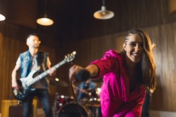 sing with us, young singer woman in pink clothes holding a microphone towards the camera and her band in the background. High quality photo