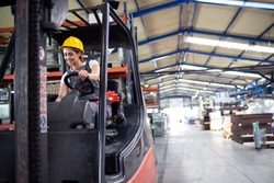 Professional female industrial driver operating forklift machine in factory's warehouse.