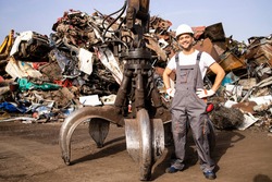 Portrait of worker standing by hydraulic industrial machine with claw attachment used for lifting scrap metal parts in junk yard.