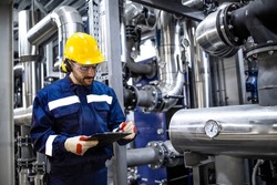 Professional worker in protective uniform and hardhat standing by pipes and checking pressure inside gasoline refinery plant.