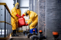 Chemical worker carrying canisters with hazardous materials and standing by large storage acid tanks.