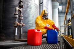 Working in chemicals production factory. Professional worker in protective hazmat suit and gas mask handling dangerous chemical next to large metal reservoirs.
