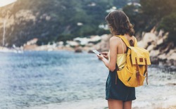 Hipster girl with backpack hold on smart phone gadget in sand coastline. Traveler using in female hand mobile on background beach seascape horizon. Tourist look on blue sun ocean, summer lifestyle 