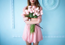 Beautiful young woman in pink dress holding spring pink flower bouquet on the blue background in the morning. 