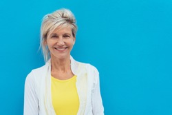 Happy vivacious older blond woman in a colorful yellow top posing against a bright blue wall with copy space