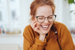 Pretty red-haired girl with pigtail, wearing glasses and orange sweatshirt, laughing with her eyes closed. Close-up front portrait indoors with copy space