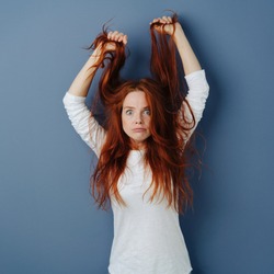 Angry young woman pulling her long red hair as she glares balefully at the camera in a rage over a blue studio background with copy space