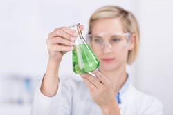 Young female laboratory technician examining a conical glass Erlenmeyer flask filled with a green liquid solution while conducting chemical tests