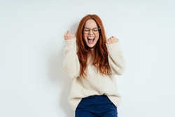 Exultant young redhead woman cheering and clenching her fists in excitement after receiving good news standing against a white wall with copy space