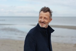 Attractive older man walking along a sandy beach on a chilly autumn day looking back over his shoulder with interest and a quiet smile