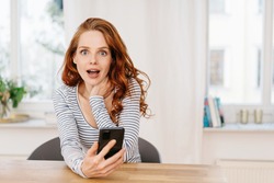 Young woman staring at camera in shocked amazement or astonishment with mouth open and wide eyes as she sits relaxing at a table in a high key room with copyspace holding a mobile phone