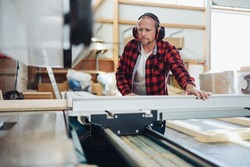 Carpenter in a woodworking workshop or factory using large industrial machinery during the production process wearing ear muffs against noise