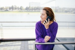 Attractive redhead middle-aged woman in trendy purple top standing looking aside waiting on hold on a call on her mobile phone on an exterior balcony with copyspace