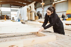 Woman worker in a carpentry workshop manufacturing prefabricated walls for houses bending down to check the alignment of a large wooden beam on a workbench