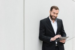 Successful businessman standing using a tablet to access the internet as he leans against a white wall with copyspace