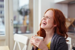 Spontaneous attractive young redhead woman enjoying a good laugh over a morning cup of coffee at home in the apartment