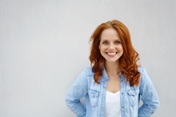 Friendly attractive young redhead student in a casual faded denim top standing against a white wall with copy space smiling at the camera