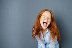 Young woman enjoying a hearty laugh leaning towards the camera with her mouth wide open over a blue studio background with copy space