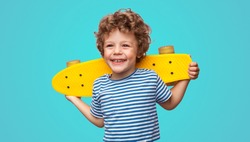 Charming curly boy holding yellow pennyboard and looking away isolated on aqua blue background.