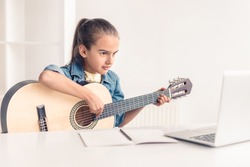 Focused little kid playing acoustic guitar and watching online course on laptop while practicing at home