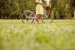 Unrecognizable female in dress standing near vintage bicycle with bouquet of flowers on green lawn on summer day in park
