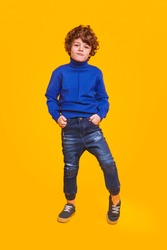 Full body cute kid in trendy sweater and jeans keeping hands in pockets and looking at camera against yellow background