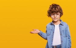 Cute child in denim shirt and glasses looking at camera and pointing at blank space isolated on yellow background