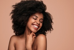 Happy African American woman with curly hair closing eyes and cheerfully smiling while enjoying clean skin after spa session against brown background