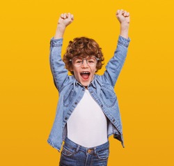 Excited schoolboy in casual wear standing with fists up on yellow background while screaming and celebrating achievement