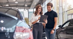 Positive young professional saleswoman demonstrating document to male customer buying new car in dealership