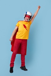 Full body excited young guy in superhero cape and helmet raising arm and screaming while pretending to fly against blue background