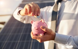 Crop unrecognizable businessman inserting coin into piggy bank while standing against blurred photovoltaic solar panel as concept of cheap alternative energy
