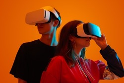Young man and woman wearing headsets of virtual reality standing on orange background looking away