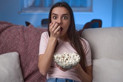 Shocked young female with long hair eating popcorn and looking at camera while sitting on sofa and watching thriller movie at night at home