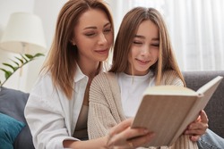 Happy mom and teen daughter smiling and reading interesting book while relaxing on couch on weekend day at home