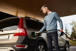 Low angle of concentrated young bearded male driver in smart casual clothes filling fuel into modern SUV car with gas pump nozzle at station
