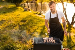 Cheerful bearded male chef in white t shirt and apron laughing and looking at camera, while cooking pork steaks and sausages on grill during picnic on lake shore on summer day