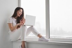 Cheerful teenager in white clothes and glasses, smiling and waving hand at start of online lesson while sitting on windowsill near window in light room at home