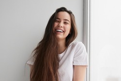 Delighted young lady with long dark hair in t shirt smiling happily and looking at camera, while sitting near window and leaning on white wall