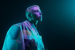 Low angle of futuristic young woman in translucent raincoat with short hair looking at colorful neon light against dark background