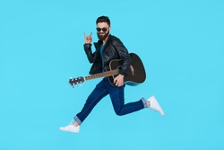 Man guitar player jumps while showing rock gesture on blue background. Isolated in motion
