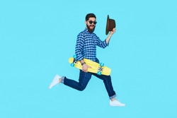 Pretty young bearded man jumping with yellow skateboard against the colorful wall. Hipster in motion on blue background