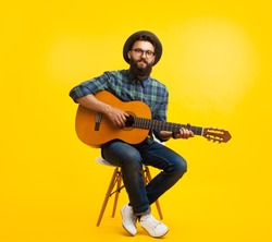 Content hipster man in hat and eyeglasses playing guitar sitting on chair on orange studio background. 