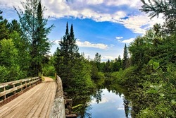 A curving wooden bridge oasses alonmgside a river and through a lush green forest on the Bearskin Trail in Northern Wisconsin.