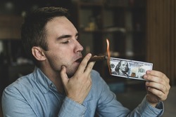 Young brutal businessman lighting cigar with 100 dollar bill as a symbol of wealth and success. The concept of wealth and extravagance.